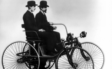 DIE ERFINDER: Daimler "Stahlradwagen" mit Zweizylinder-V-Motor und dem ersten Zahnrad-Schaltgetriebe samt Differentialausgleich, vorgestellt von Gottlieb Daimler und Wilhelm Maybach (am Steuer) auf der Weltausstellung in Paris 1889 - Daimler AG